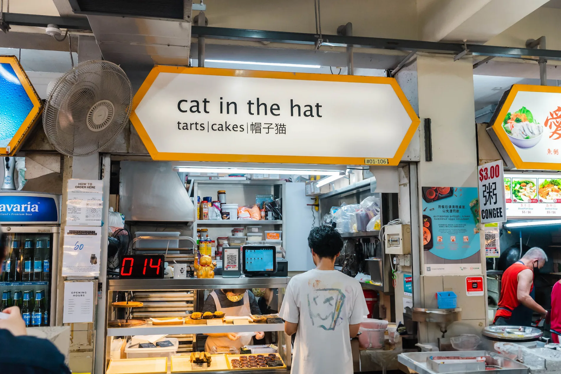 The cat in the hat stall at Golden Mile Food Centre, unit #01-106, its yellow-framed signboard reading tarts, cakes, 帽子猫 above the display counter and the red queue-number display below